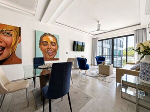 A modern living room with a glass dining table, blue chairs, a sofa, and a large window with a view of buildings. - Stylish 1-Bed – LAP Pool - Wi-Fi - GYM - Hot water (Sosúa)