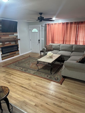 A living room with a wooden floor, a gray sofa, a coffee table, a ceiling fan, and a fireplace. - The Phoggy Cottage (Lawrence)