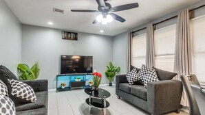 A modern living room with a ceiling fan, a flat-screen TV, a sofa, a coffee table, and potted plants. - Modern Cozy OKC Stay Near Fairgrounds (Oklahoma City)