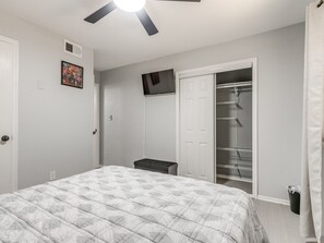 Une chambre à coucher comprenant un lit, une armoire avec des étagères, un téléviseur fixé au mur et un ventilateur de plafond.