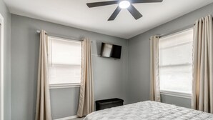 A bedroom with a bed, a ceiling fan, a TV mounted on the wall, and two windows with curtains. - Modern Cozy OKC Stay Near Fairgrounds (Oklahoma City)