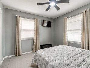 Ein Schlafzimmer mit einem Bett, einem Deckenventilator, einem an der Wand montierten Fernseher und zwei fensterhohen Vorhängen.