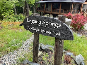 Ein Holzschild für den Legacy Springs Trail, umgeben von Grünpflanzen und einem Gebäude im Hintergrund.