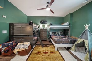Ein Schlafzimmer mit Etagenbetten, einem Schreibtisch, einem Stuhl, einem Bett mit gemustertem Bettbezug, einem kleinen Zelt und einem Deckenventilator.