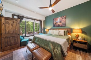 Quarto com uma cama grande, um banco, um ventilador de teto e uma janela com vista para árvores.