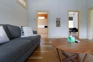 Ein Wohnzimmer mit grauem Sofa, einem runden Holztisch und einem durch eine offene Tür einsehbaren Küchenbereich.