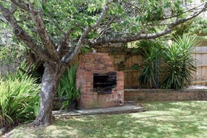 Un barbecue extérieur en briques, doté d’une cheminée, entouré d’une cour verdoyante et d’une clôture en bois.