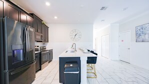 A modern kitchen with dark cabinetry, stainless steel appliances, a central island with bar stools, and a wall-mounted clock. - Sweet Home Near Disney —5BR Resort Villa + Private Pool (Kissimmee)