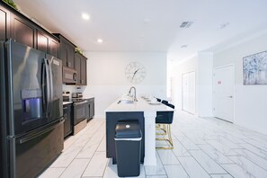 A modern kitchen with dark cabinetry, stainless steel appliances, a central island with bar stools, and a wall-mounted clock. - Sweet Home Near Disney —5BR Resort Villa + Private Pool (Kissimmee)