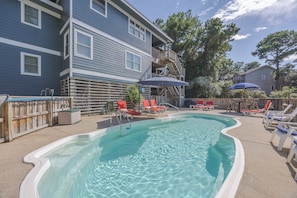 A swimming pool with lounge chairs and umbrellas, surrounded by a wooden deck and a building with a balcony. - 1062 - Beach House Blessings Spacious 5BR Corolla retreat with private pool, deck, and elevator. (Corolla)