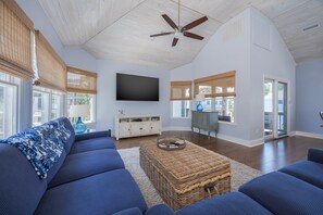 A spacious living room with a large flat-screen TV, a ceiling fan, and a view of the outdoors through large windows. - 1062 - Beach House Blessings Spacious 5BR Corolla retreat with private pool, deck, and elevator. (Corolla)