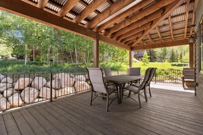 Eine überdachte Terrasse im Freien mit einem Holztisch und -stühlen, umgeben von einem Metallgeländer und Blick auf ein bewaldetes Gebiet.