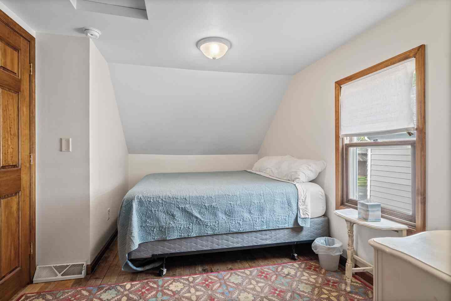 A small, single-bed room with a sloped ceiling, a window with blinds, a radiator, and a wooden door.