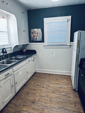 A kitchen with white cabinets, a wooden floor, a refrigerator, and a window with blinds.