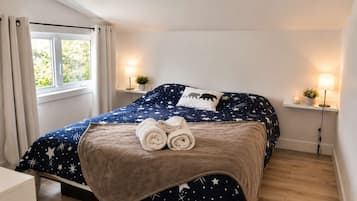 Une chambre à coucher avec un lit, deux lampes de chevet, une fenêtre avec des rideaux et une table de chevet ornée d’une plante.
