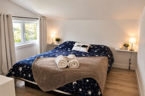 Un dormitorio con cama, dos lámparas de noche, una ventana con cortinas y una mesita de noche con una planta.
