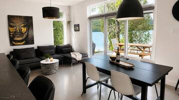 Un espace de vie moderne avec un canapé noir, une table à manger avec des chaises et une grande fenêtre donnant sur une étendue d’eau.