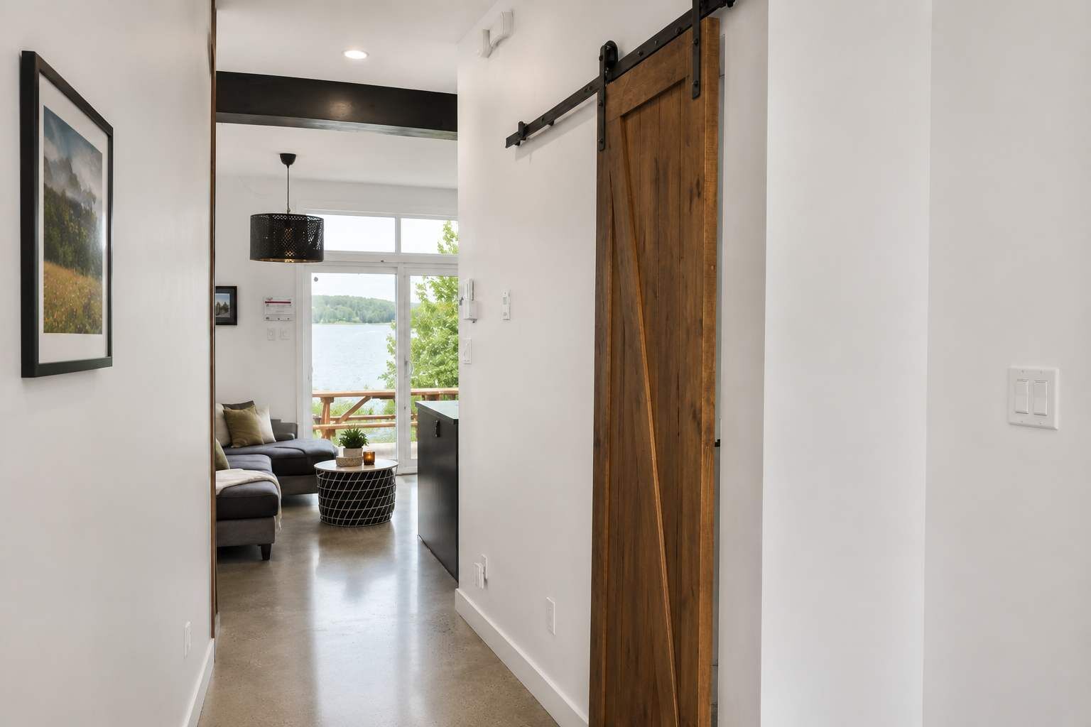 Un couloir doté d’une porte coulissante de style rustique, d’un canapé, d’une table basse ronde et d’une vue sur un lac, visible à travers une porte en verre.