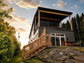 A modern two-story house with a wooden deck, a staircase leading to the entrance, and a large window.