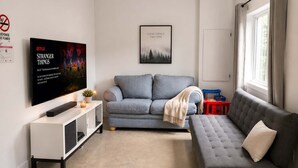 Una sala de estar con un sofá gris, un sofá a rayas, un televisor de pantalla plana, un cuadro enmarcado y una ventana con cortinas.