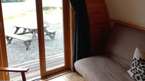 A cozy room with a wooden ceiling, a sofa with a patterned pillow, and a view of an outdoor picnic area through a glass door.