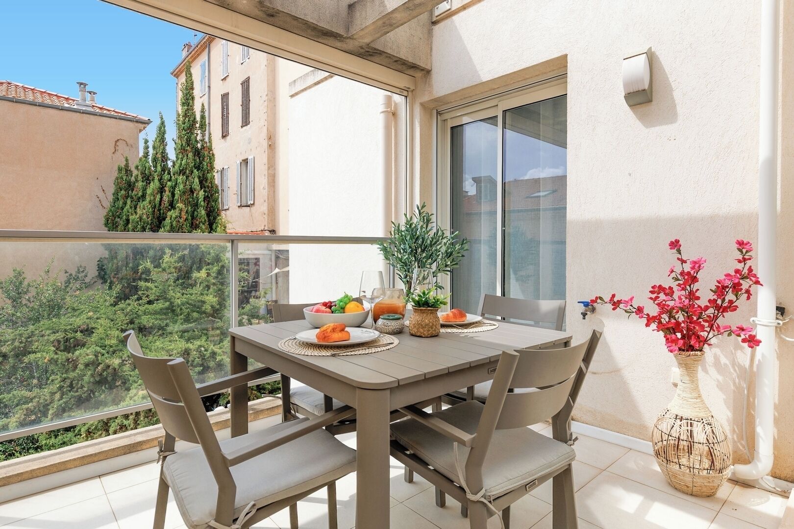 A balcony with a table set for two, featuring a bowl of fruit, a glass of juice, and a potted plant.