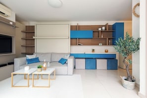 Ein modernes Wohnzimmer mit grauem Sofa, einem Couchtisch und einem Fernseher. Es gibt eine Küchenzeile mit blauen Schränken und einer Pflanze.