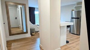 Un vaste espace de vie doté d’un plancher en bois, d’un mur blanc et d’une porte-fenêtre donnant sur une cuisine équipée d’un réfrigérateur en acier inoxydable.