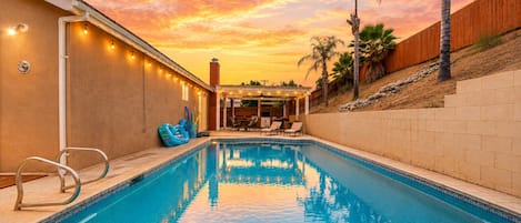 A swimming pool with a sunset view, a covered patio area, and a wooden fence.