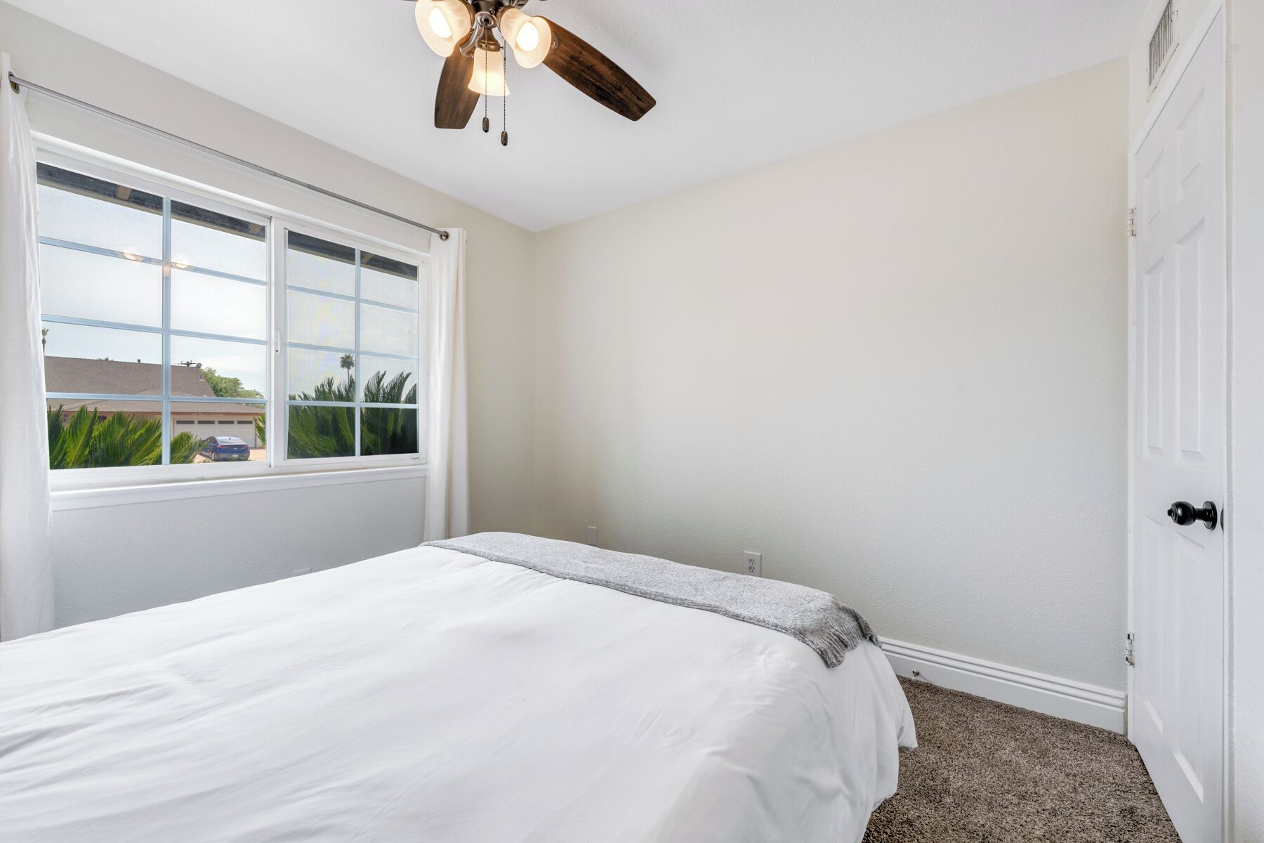 Une chambre à coucher avec un lit, un ventilateur de plafond, une fenêtre donnant sur une maison et des palmiers, et une porte.