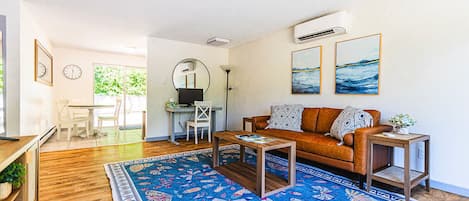 A living room with a sofa, coffee table, and a rug. There is a desk with a computer and a chair. The room has a large window and an air conditioning unit on the wall.
