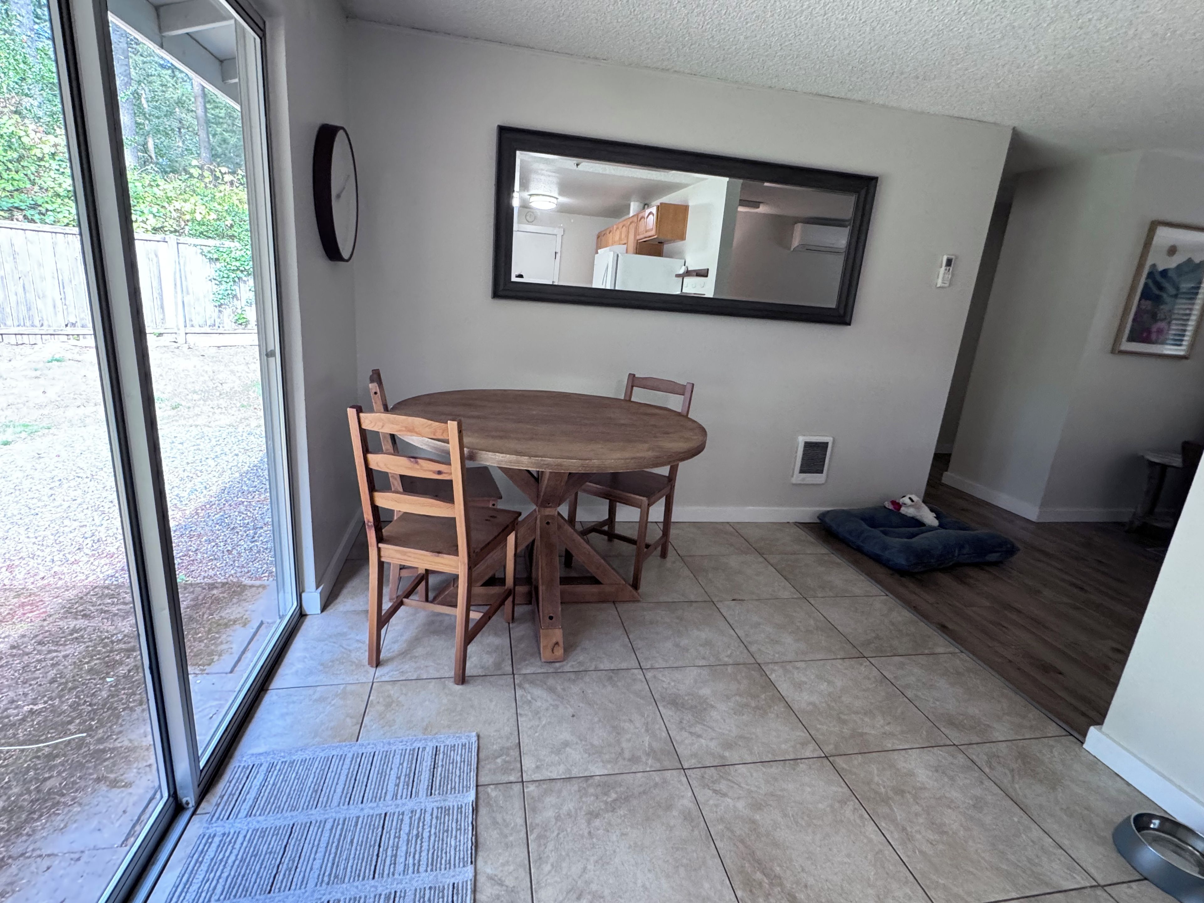 A dining area with a round table and chairs, a large mirror on the wall, and a pet bed in the corner.