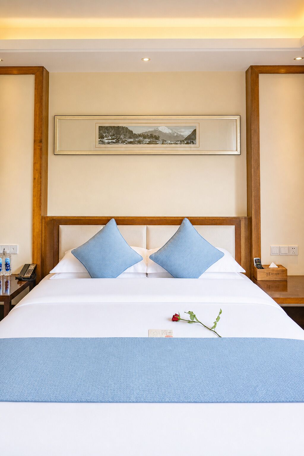 A hotel room with a large bed, two pillows, a bedside table with a phone, a remote control, and a small box.