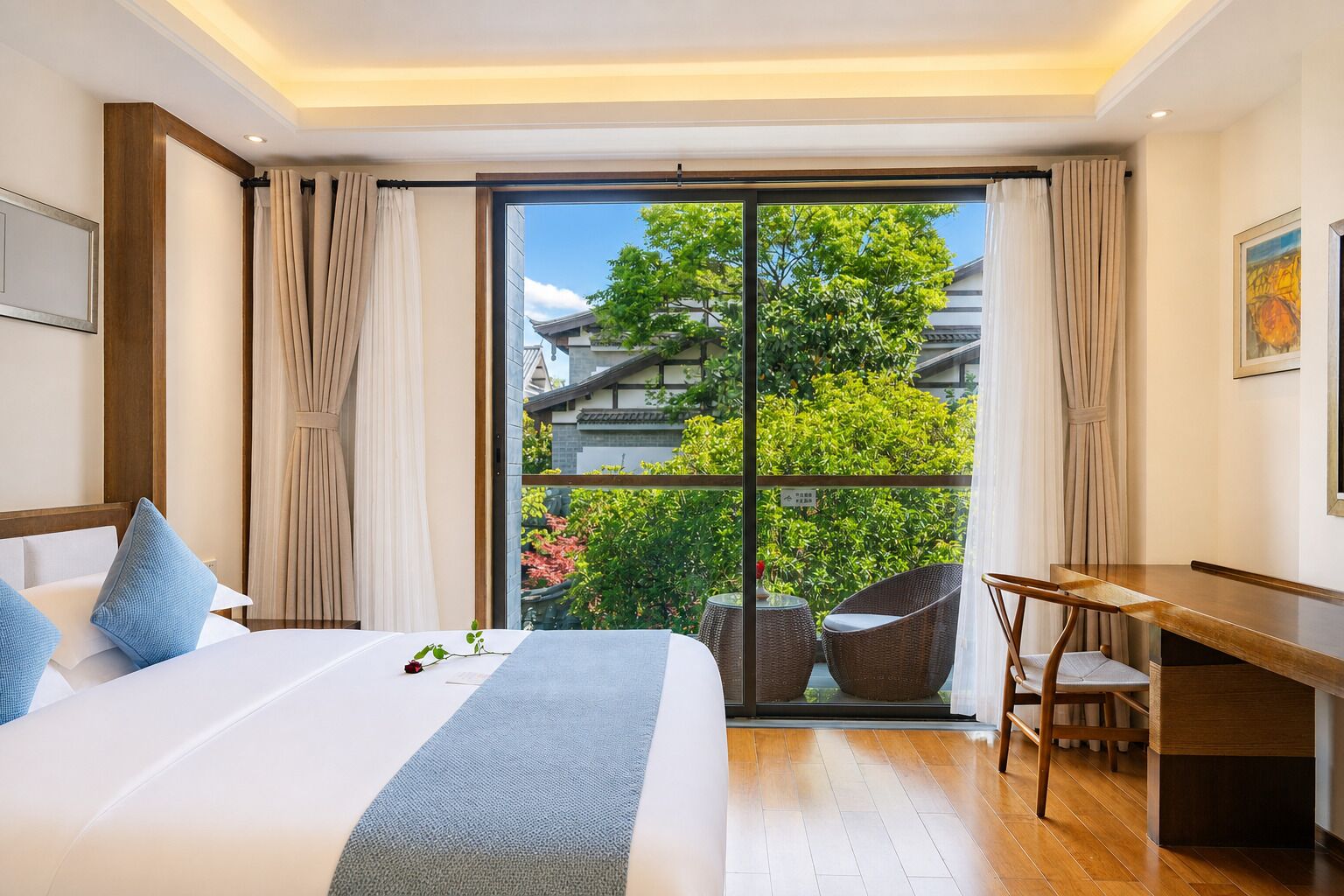 Ein Hotelzimmer mit einem großen Bett, einem Schreibtisch und Blick auf ein Gebäude und Bäume durch eine Glastür.