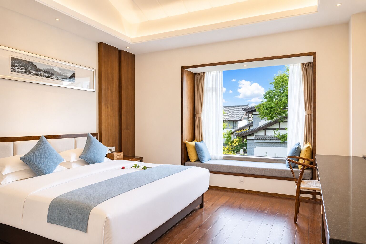 Habitación de hotel con una cama grande, cabecera de madera, un banco junto a la ventana con vistas a un edificio y árboles, y un escritorio con una silla.