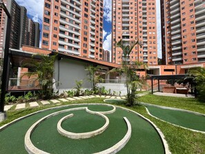 Zona residencial con edificios altos, un jardín ajardinado con césped artificial verde y un área de estar al aire libre techada.