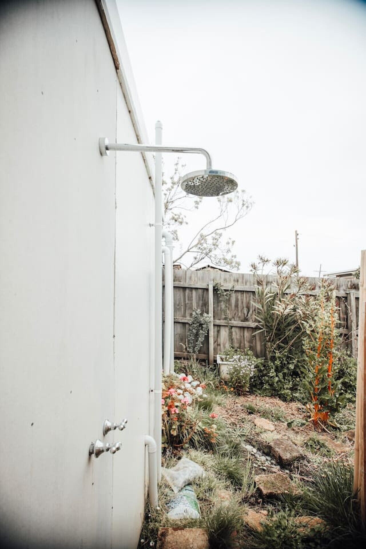 A metal structure with a showerhead, a wooden fence, and various plants in a garden area.