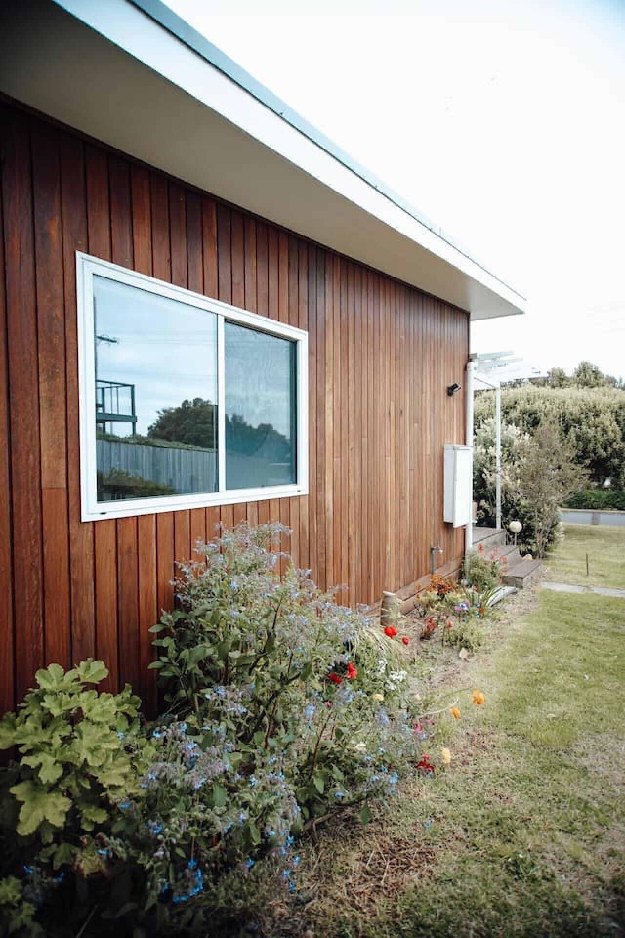 A modern building with a wooden exterior, a large window, and a well-maintained garden with various plants and flowers.