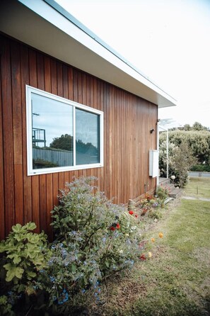 A modern building with a wooden exterior, a large window, and a well-maintained garden with various plants and flowers. - Craypot Cottage - Port Campbell, Victoria (Port Campbell)