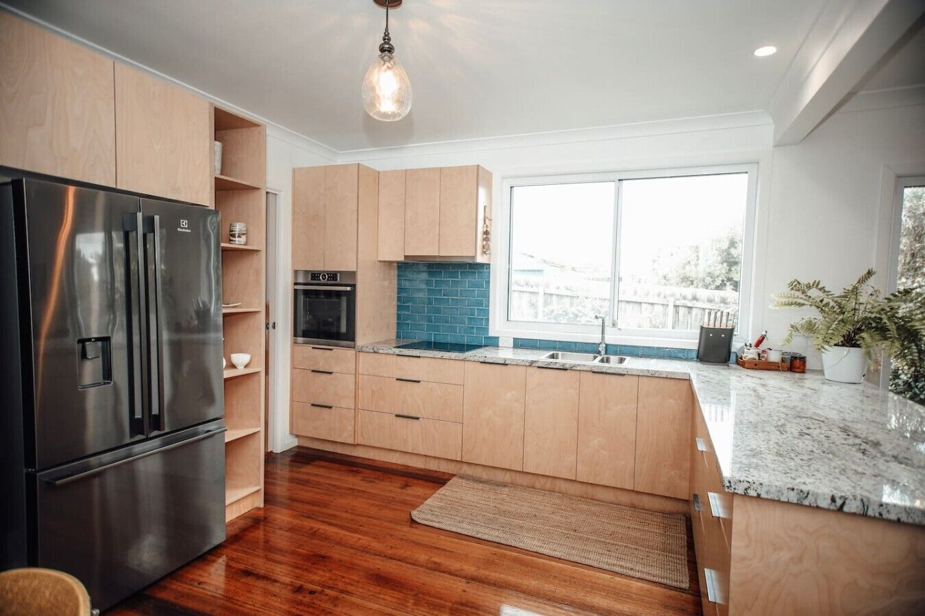A modern kitchen with wooden cabinets, a stainless steel refrigerator, a built-in oven, and a large window providing a view.