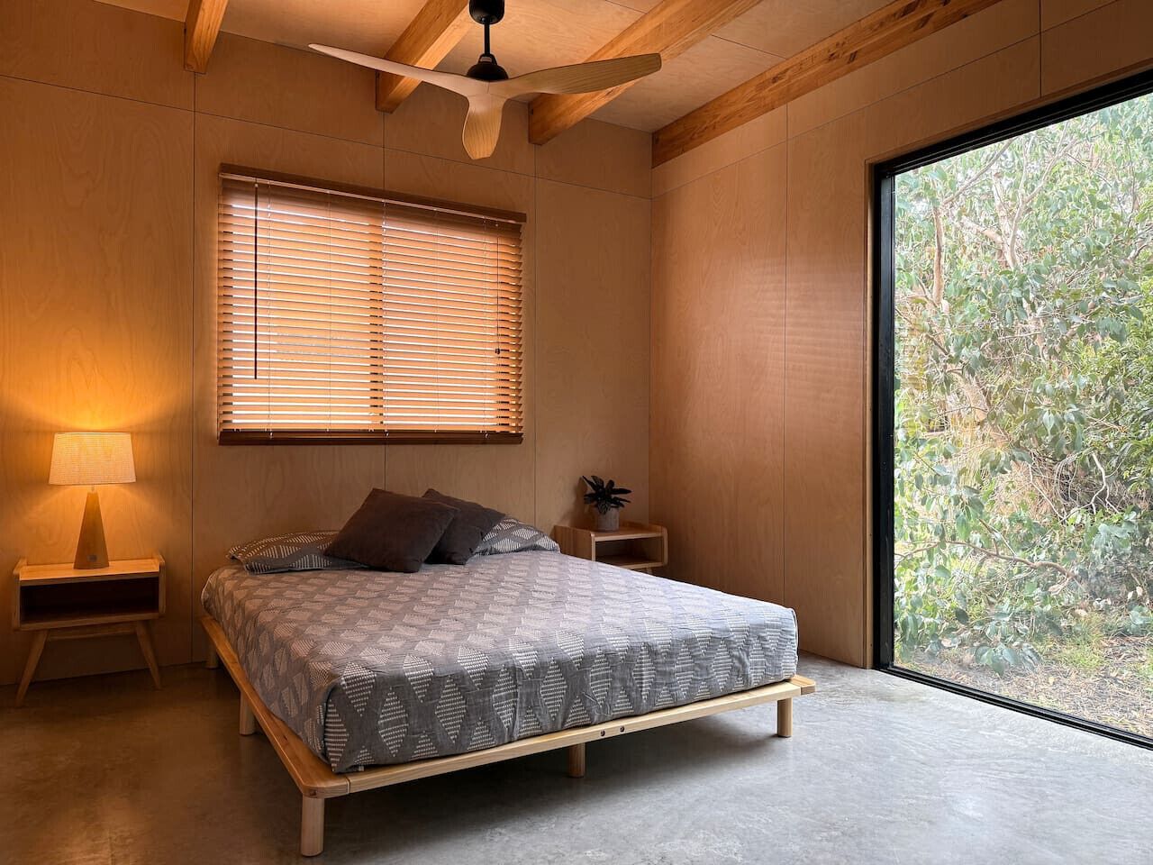 Une chambre à coucher comprenant un lit, une table de chevet, une lampe, un ventilateur de plafond et une grande fenêtre avec des stores.