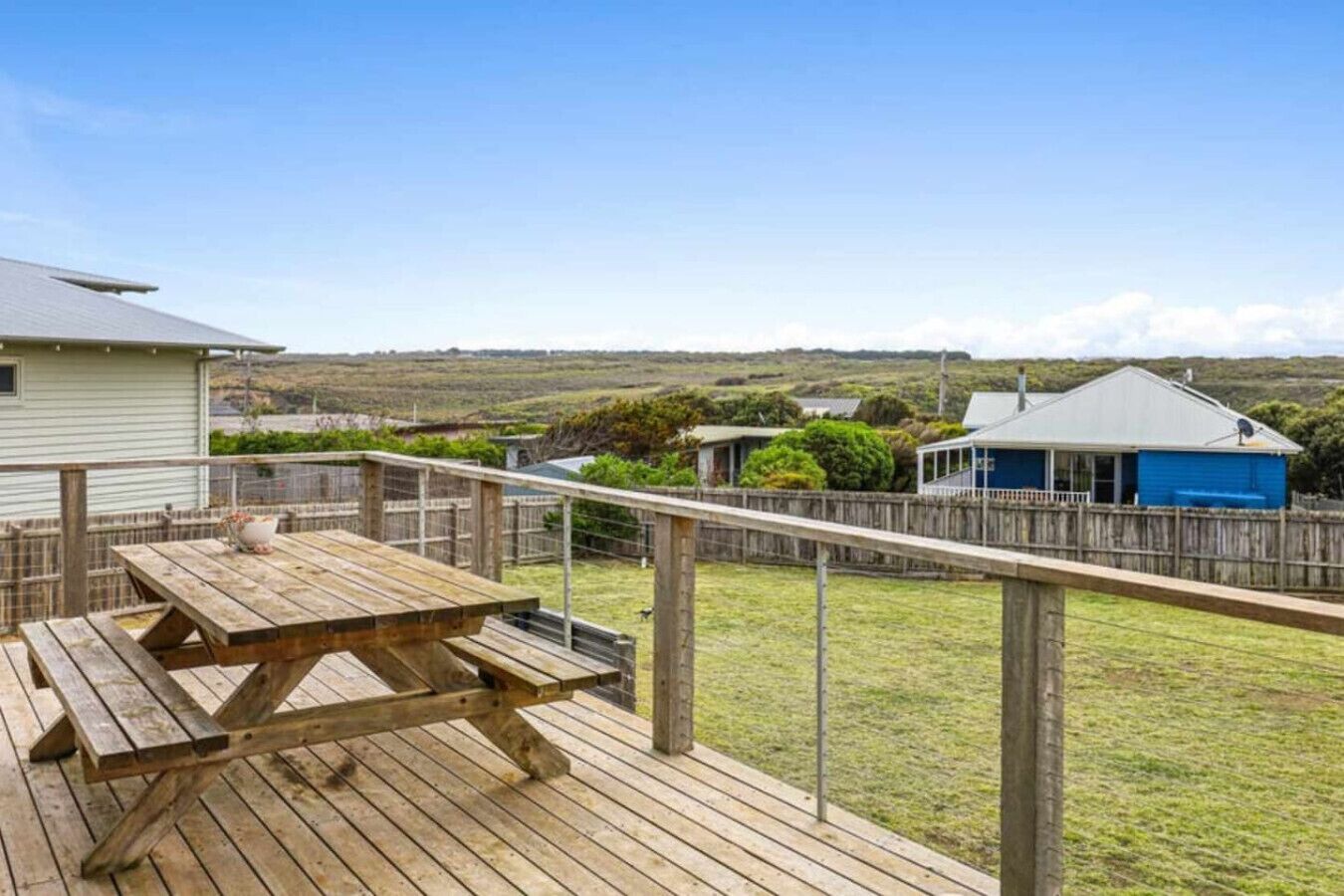 Un terrazzo in legno con un tavolo da picnic, circondato da una recinzione di legno, con vista su una zona erbosa e un edificio blu sullo sfondo.
