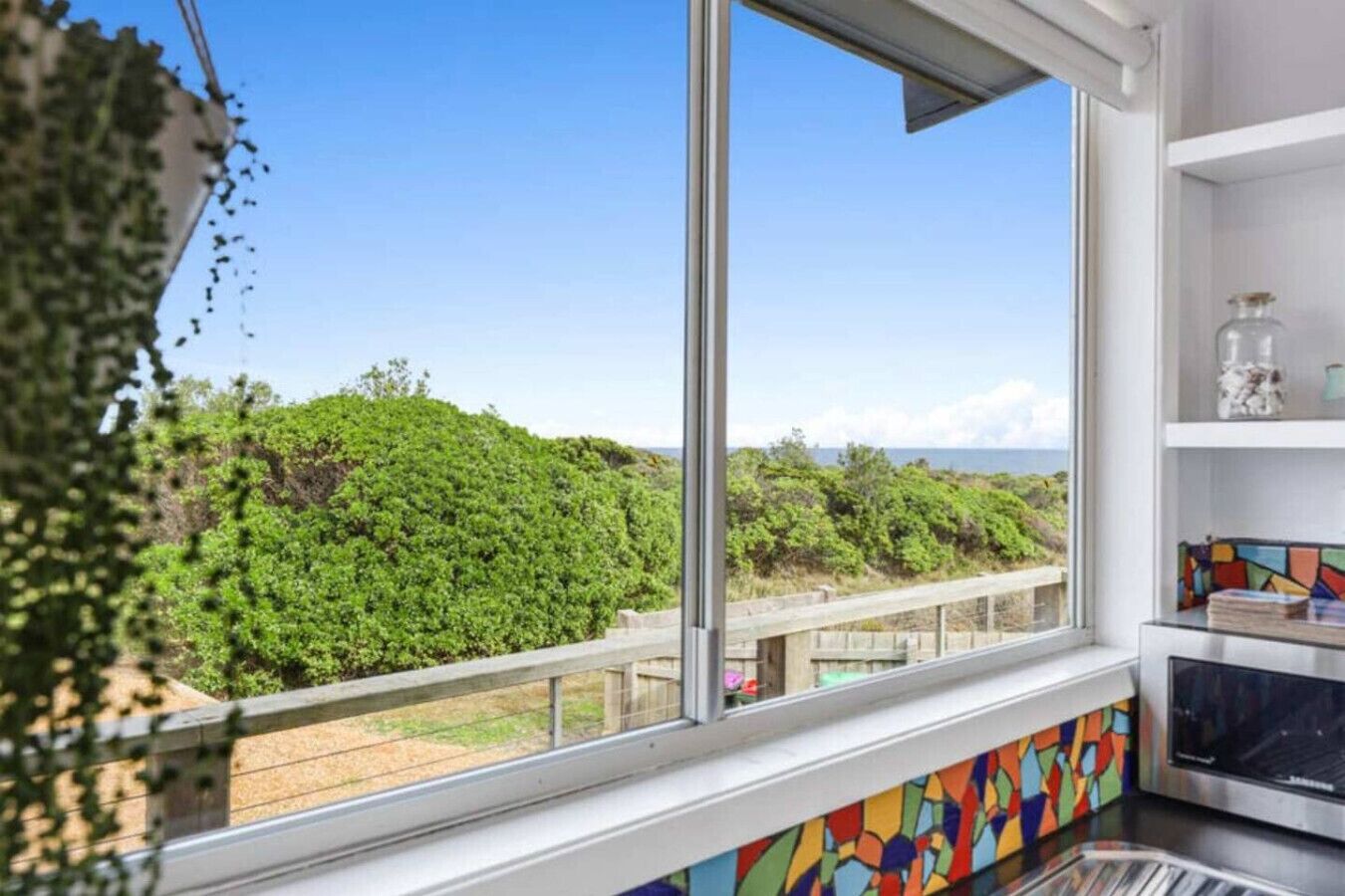 A room with a large window overlooking a coastal landscape, a kitchen area with a colorful backsplash, and a view of the sea.