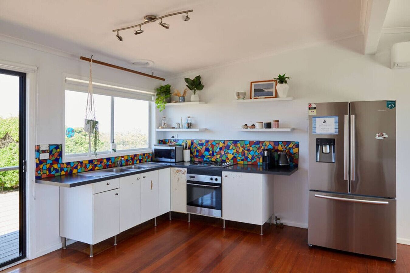 Una cucina moderna con mobili bianchi, un backsplash colorato, un frigorifero in acciaio inossidabile e una finestra con una pianta sospesa.
