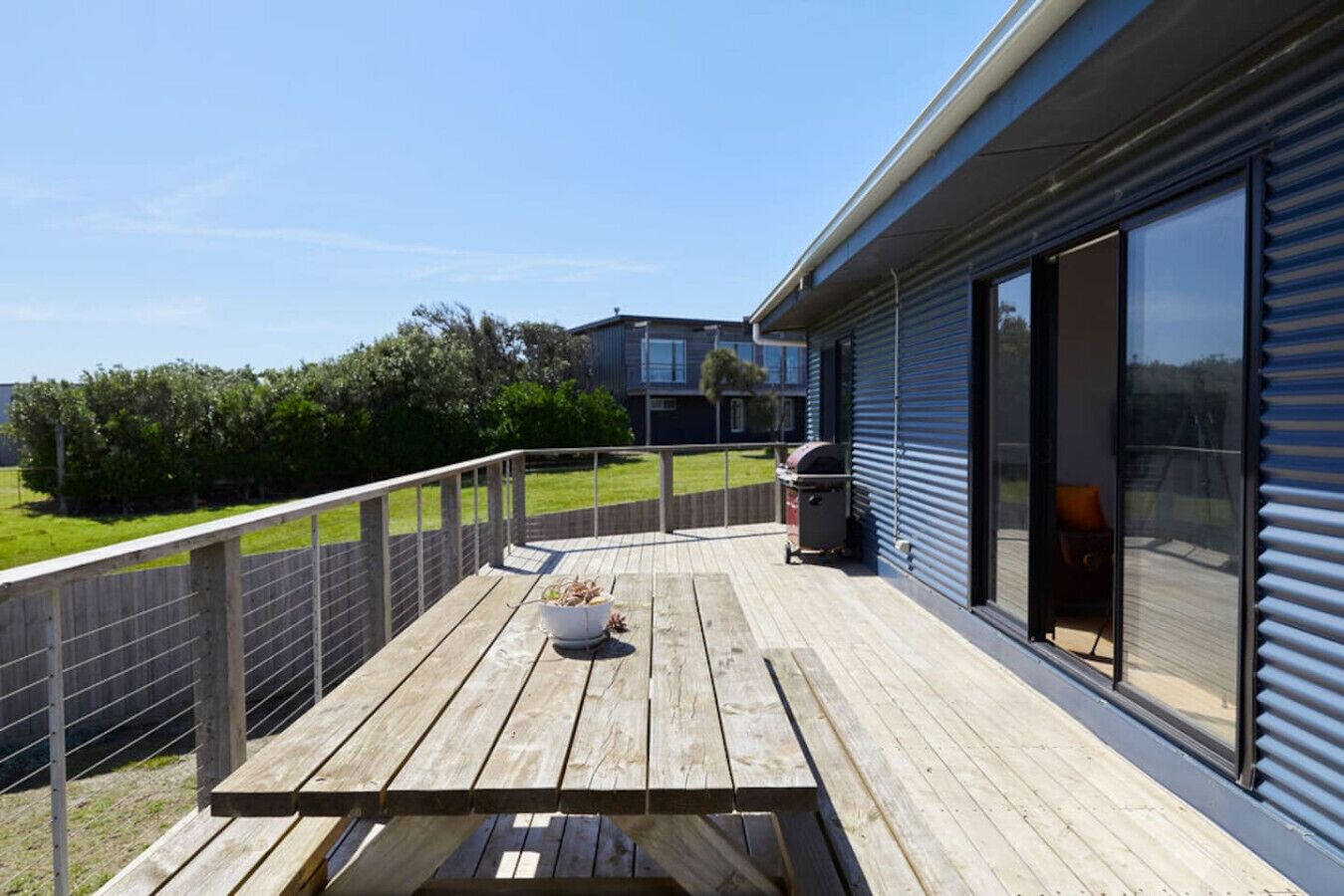 Un terrazzo in legno con una griglia per barbecue e una pianta in vaso, con vista su una zona verde e un edificio con ampie finestre.