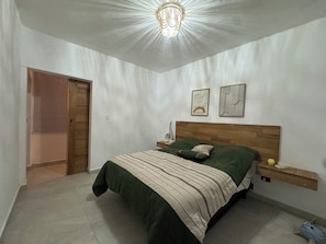 A bedroom with a bed, a wooden headboard, a nightstand, a wall-mounted shelf, and a door.