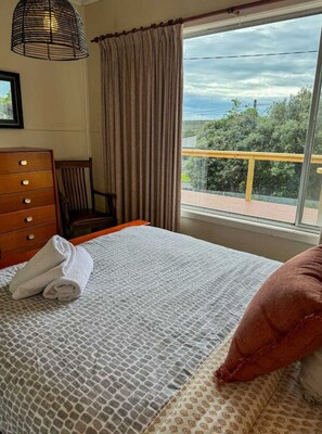 Una cama con una colcha estampada, una toalla enrollada y un almohilar rojo. Se observa un aparador de madera con cajones y una silla. Una ventana con cortinas muestra una vista de follaje y una terraza de madera al exterior.