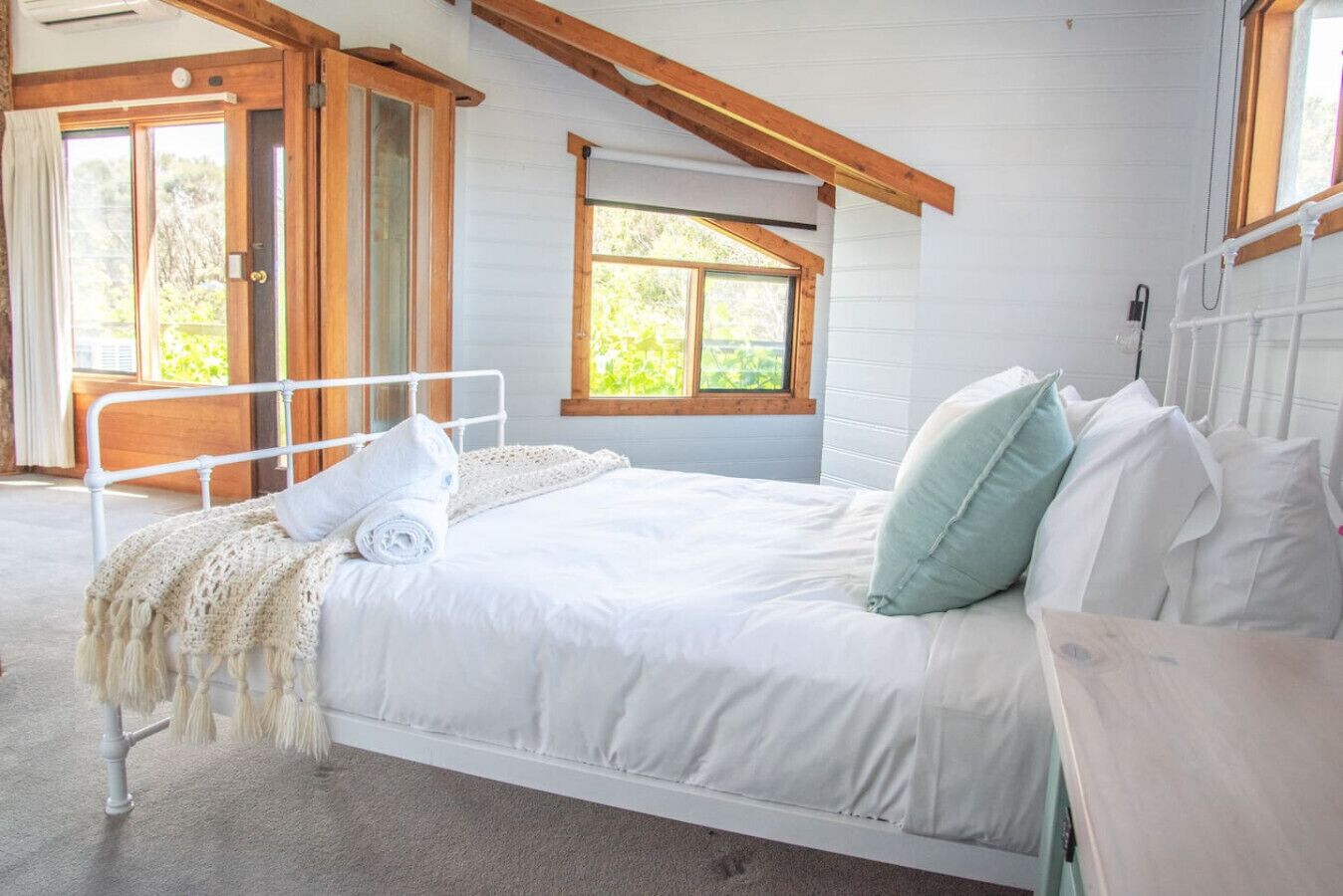Ein Schlafzimmer mit weißem Metallbettgestell, einem aufgerollten weißen Handtuch und einem hellblauen Kopfkissen. Eine Holztür führt nach draußen, und ein Fenster bietet Blick auf Grünflächen.