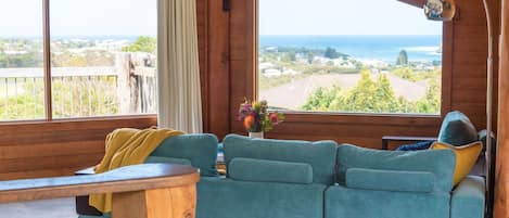 A room with a large window offering a view of the sea and landscape, a teal sofa with cushions, and a wooden table.