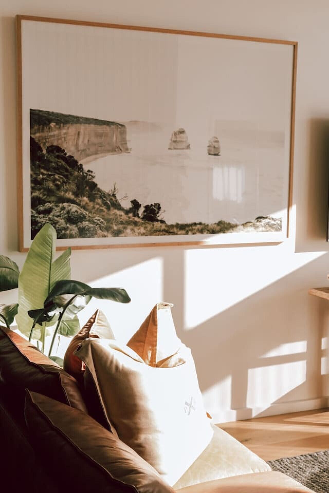 A room with a large framed artwork on the wall, a brown leather sofa, a white pillow, and a plant.